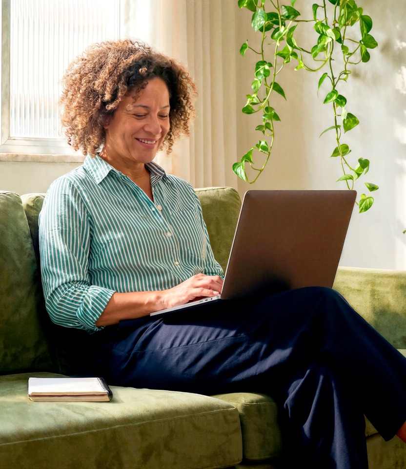 Mulher madura sorridente sentada no sofá de casa usando o notebook