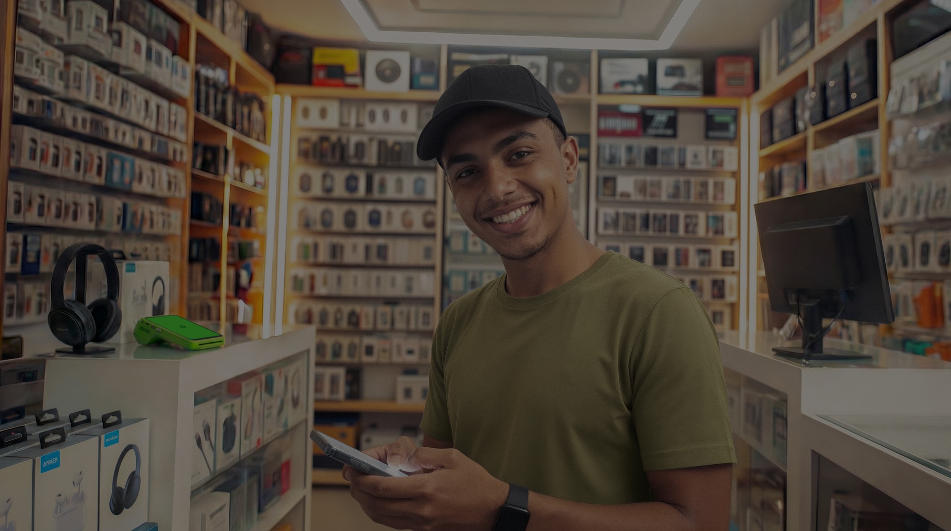 Empreendedor sorridente em loja de acessórios eletrônicos segurando o celular, com fones e produtos expostos ao fundo
